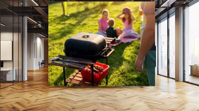 Man is about to use a vintage old clean grill with meat and vegetables already ready beneath it while kids are chatting and sitting on the blanket that has been put on grass. Wall mural