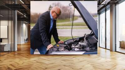 Mature professional elegant worried businessman in the suit is looking under the car hood trying to figure out the problem while leaning against the car. Wall mural