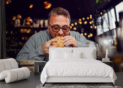 Middle aged bearded hungry man sitting in restaurant and eating delicious burger. Wall mural