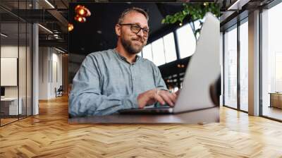 Middle aged bearded smiling freelancer with eyeglasses sitting in a restaurant and using laptop to type report. Middle aged persons can be excellent freelancers. Wall mural