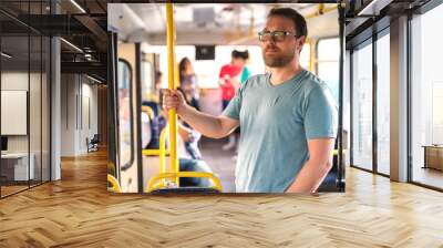 Middle-aged Caucasian man driving in the city bus while standing and holding grip. In background people sitting and standing. Wall mural