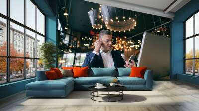 Middle-aged Caucasian man with serious face dressed smart casual using smart phone for call while sitting in cafe. In the desk laptop and coffee. Wall mural