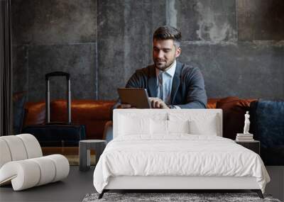 Middle-aged handsome businessman sitting on a couch in a hotel lobby and check his online social networks on a digital tablet. Next to him is luggage. Business trip, travel, technologies Wall mural