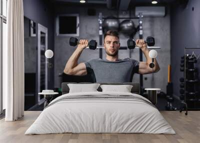 Muscular guy lifting dumbbell while sitting on a bench at the gym. A young athlete using dumbbells during a workout. Strong man under physical exertion pumping up bicep muscle with weight Wall mural