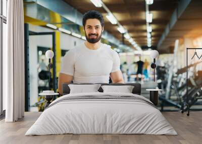 Muscular handsome bearded trainer looking at the camera while standing with a tablet in the gym. Wall mural