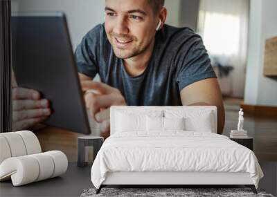 Online tablet research. A happy male person lies on the floor in the room and checks the news in the world. He uses a tablet and scrolls the touch screen with his finger. Next to it is a telephone Wall mural