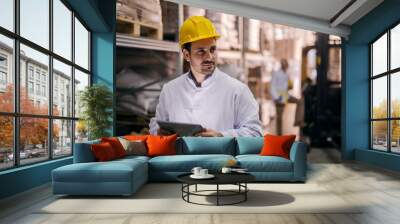 Picture of young focused male manager standing in factory and holding tablet. Dressed in white coat with helmet on his head. Looking satisfied and proud. Wall mural