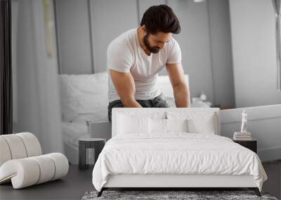 Portrait of unhappy handsome muscular bearded man having knee pain and sitting on the edge of the bed at home. Wall mural