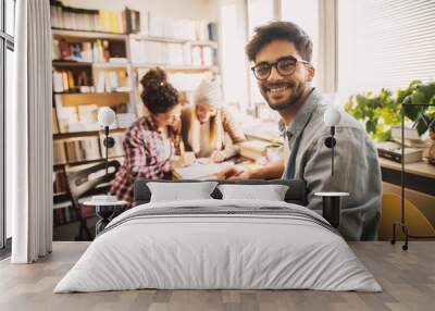 Portrait view of handsome nerdy smart young man with glasses doing homework with two beautiful girls sitting in the background in the school library. Wall mural