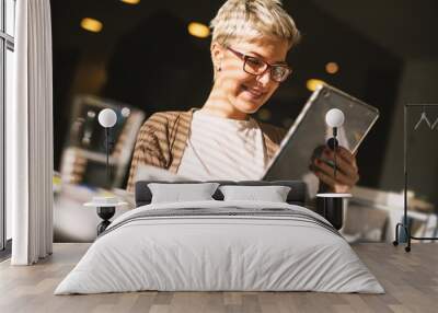 Pretty girl with glasses and short hair checking the news on a tablet while drinking a coffee. Wall mural