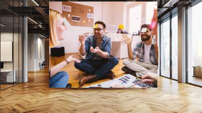 Pretty young designer coworkers enjoying free time and raising togetherness and team spirit while sitting in a circle on the floor of the office and playing games with paper stickers on the forehead. Wall mural