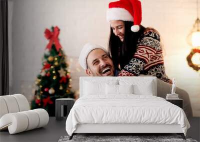 Romantic young couple enjoying Christmas time. Handsome smiling man giving piggyback to his wife at home for Christmas holidays. Wall mural