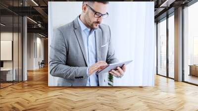 Serious caucasian classy businessman in suit and with eyeglasses standing next to window and using tablet at his office. Wall mural