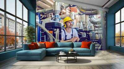 Serious caucasian handsome worker in overall, with hardhat and antiphons crouching and using tablet. Factory interior. Wall mural