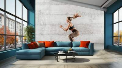 Side view of muscular focused brunette with ponytail and in sportswear jumping in front of brick wall in gym. Wall mural