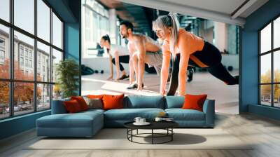 Small group of people with healthy habits doing stretching exercises on a gym floor. Selective focus on blonde woman. In background mirror. Wall mural