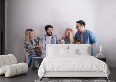 Small group of young businesspeople holding coffee to go and smart phones while talking and laughing. In background while wall. Start up business concept. Wall mural