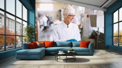 Smiley female worker in sterile clothes using tablet and checking how production line is working. Wall mural