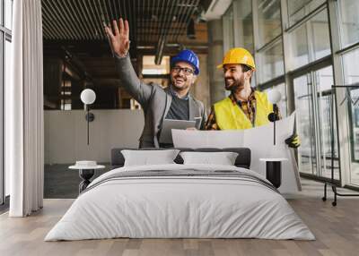 Smiling architect holding tablet and showing to construction worker something he imagined. Construction worker holding blueprints. Construction site interior. Wall mural