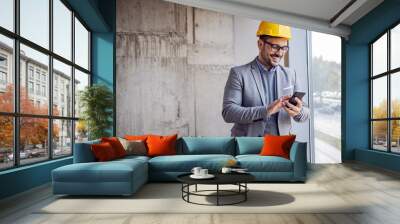 Smiling architect in suit and with helmet on head standing in building in construction process next to window and typing message on smart phone. Wall mural