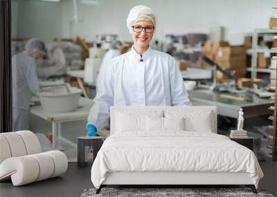 Smiling blonde Caucasian employee in sterile uniform and with eyeglasses standing and holding tray with cookies. Food factory interior. In background other employees working. Wall mural