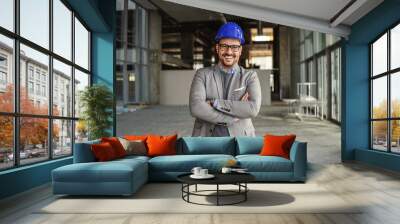 Smiling businessman with helmet on head standing with arms crossed in his building in construction process and looking at camera. Wall mural