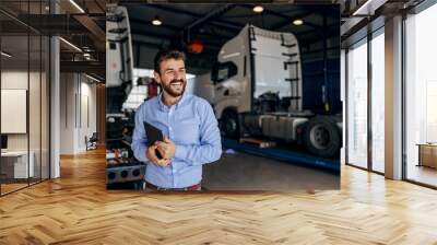 Smiling chief standing in auto park and holding tablet. In background are trucks. Firm for import and export. Wall mural