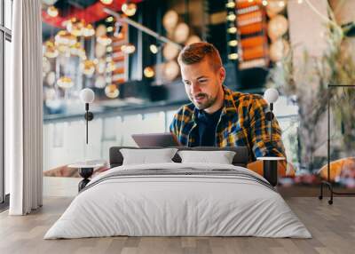 Smiling freelancer using tablet for work. In front of him on a table cup of coffee. Cafeteria interior. Wall mural