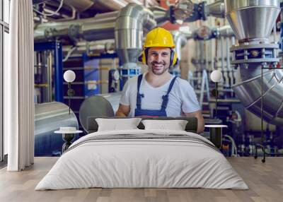 Smiling handsome caucasian blue collar worker in overall, with hardhat and antiphons holding tablet while standing in factory and looking at camera. Wall mural