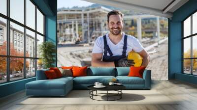 Smiling handsome unshaven caucasian worker in overall standing at refinery with helmet under armpit and looking at camera. Wall mural