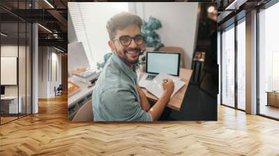 Smiling mixed race male student with eyeglasses doing school project by using laptop and looking at camera. Wall mural