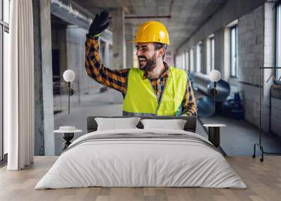 Smiling positive construction worker in work wear standing in building and waving to his colleague. Building in construction process interior. Wall mural