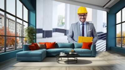 Smiling satisfied caucasian in gray suit with helmet on head and folder in hands posing outside. Wall mural