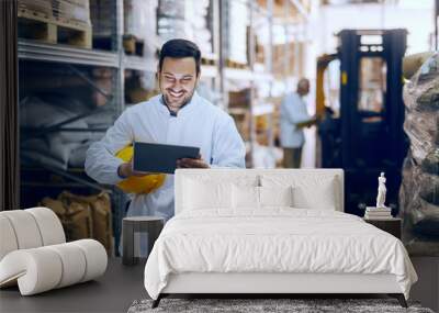 Smiling young Caucasian blue collar worker in white uniform and with helmet under armpit using tablet for controling delivery. In background worker standing next to forklift. Warehouse interior. Wall mural