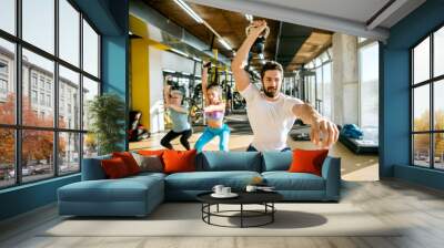 Strong muscular handsome young personal trainer doing exercises with kettlebells with two active sporty fitness women in the sunny modern gym. Wall mural