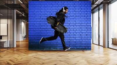 stylish young dreadlocks hipster with headphones walking with a skate in hand near the blue brick wa Wall mural