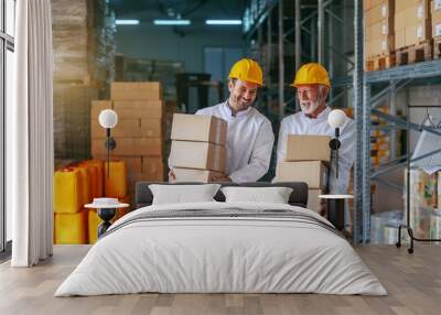 Three quarter length of two smiling Caucasian storage employees in white uniforms and with yellow helmets carrying boxes in storage. Wall mural