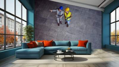 Top view of supervisor with helmet on head in suit standing with construction worker, holding tablet and explaining him ideas about project. Wall mural