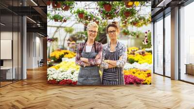 Two cheerful happy attractive middle-aged florist women posing in a large greenhouse full of colourful different flowers. Wall mural