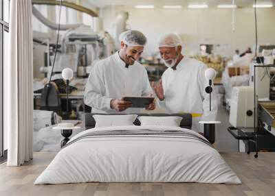 Two cheerful male colleagues in sterile clothes standing in food factory and looking at sales documents on digital tablet. Wall mural