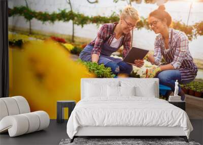 Two cheerful pretty curious florist girls having a conversation in the sunny greenhouse full of colourful flowers while kneeling and watching tutorials from a tablet. Wall mural