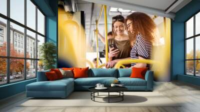 two cheerful pretty young women are standing in a bus and looking at the phone and smiling while wai Wall mural