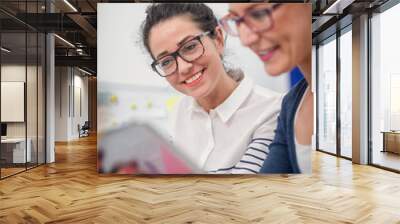 Two happy beautiful businesswoman sitting in their office and looking at tablet. Wall mural