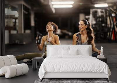 Two happy sporty friends sitting on a gym floor and drinking water while taking a break. Wall mural