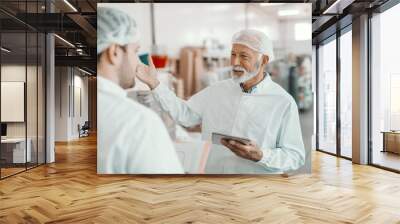 Two supervisors discussing about quality of food in food plant. Younger one holding folder with data while older one holding tablet. Both are dressed in white sterile uniforms and having hairnet. Wall mural