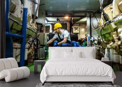 Worthy caucasian mechanic in overalls and with helmet kneeling inside ship and repairing engine. Wall mural