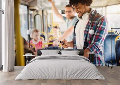 Young Afro-American with headset around his neck is looking at his phone while riding in a bus. Wall mural