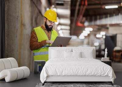 Young attractive bearded supervisor standing inside of garage in construction process and checking on an e-mail on laptop. Wall mural