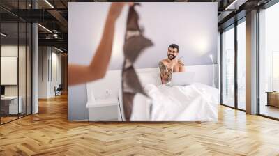 young attractive caucasian bearded tattooed man sitting on bed in bedroom with laptop in lap and loo Wall mural