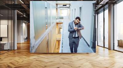 Young bearded businessman looking at wristwatch and talking with business partners. He is late for meeting and making excuse. Building in construction process interior. Wall mural